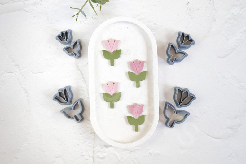 May include: A set of five cookie cutters in the shape of flowers. The cutters are made of a light gray plastic and are arranged on a white surface. The flowers have pink petals and green stems.