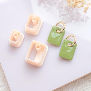 Puede incluir: Un conjunto de suministros para manualidades, que incluye tres cortadores de arcilla polim&eacute;rica de color melocot&oacute;n claro en forma rectangular y dos pendientes verdes con aros dorados. Los pendientes tienen un acabado brillante.