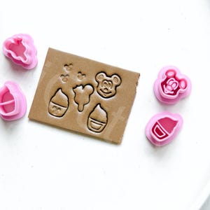 Peut inclure: Un ensemble d'emporte-pièces en plastique rose avec différents motifs, dont un ours, un cupcake et une bouteille. Les emporte-pièces sont disposés sur une surface blanche à côté d'un morceau de papier brun sur lequel les mêmes motifs sont en relief.