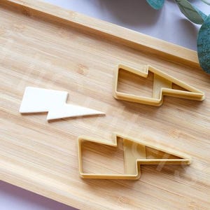 May include: A white lightning bolt-shaped cookie cutter and two yellow lightning bolt-shaped cookie cutters are displayed on a wooden tray. The cutters are for baking and crafting. The tray has a light brown color.