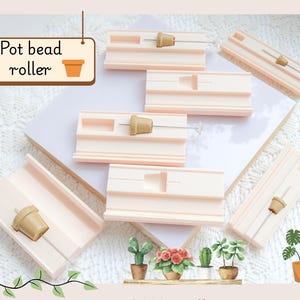 Può includere: Una collezione di rulli per perline a forma di vaso rosa chiaro con piccole perline di legno a forma di vaso. L'immagine include un cartello con la scritta "Pot bead roller" e una grafica di vaso. Sono visibili anche piante decorative.
