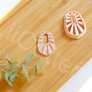 Könnte beinhalten: Zwei hellbraune Ohrringe aus Ton mit geometrischem Design. Die Ohrringe liegen auf einer Holzoberfläche.