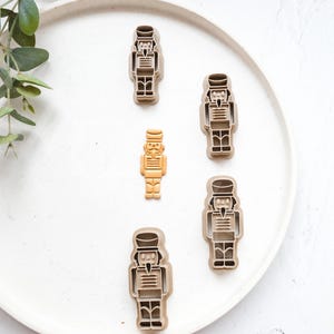 May include: Five brown plastic nutcracker cookie cutters on a white plate. One of the nutcracker cookie cutters is gold.