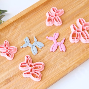 Könnte beinhalten: Ein Set mit sechs rosafarbenen und grauen Plastik-Ausstechern in Form von Ballontieren. Die Ausstecher sind auf einer Holzoberfläche angeordnet.