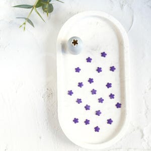 Può includere: Un vassoio ovale bianco con un piccolo vaso grigio e spolverata di fiori viola.