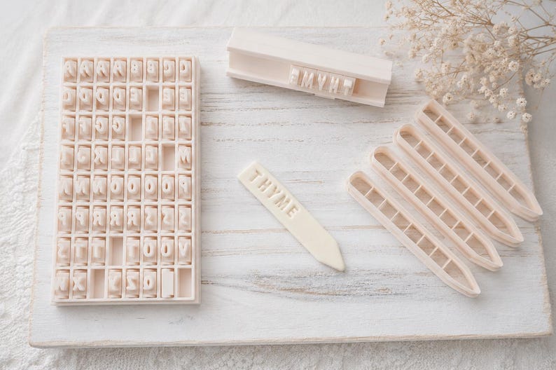 May include: A set of off-white plastic tools for clay or dough, including a rectangular grid tray filled with individual letter stamps, a long rectangular stamp holder with the word THYME, four pointed plant markers, and one marker labeled THYME.