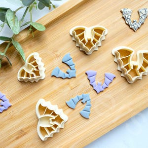 Puede incluir: Un conjunto de ocho cortadores de galletas con forma de murciélagos. Los cortadores están hechos de plástico blanco y están dispuestos sobre una superficie de madera. Hay dos tamaños diferentes de cortadores de murciélagos, y algunos de los cortadores están apilados uno encima del otro.