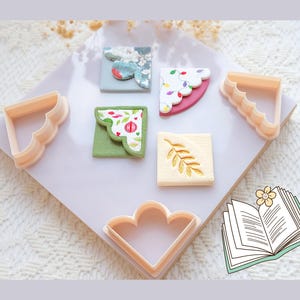 Puede incluir: Vista aérea de cortadores de galletas color melocotón y galletas decoradas. Los cortadores tienen un borde festoneado. Las galletas cuadradas están decoradas con diseños florales y abstractos. También hay una pequeña ilustración de un libro abierto con una flor.