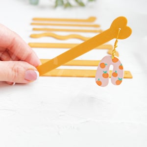 Może przedstawiać: Para różowych i pomarańczowych kolczyków z pozłacanym haczykiem. Kolczyki mają kształt łuku i wzór z małych pomarańczowych kółek z zielonymi liśćmi.