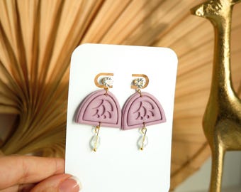 Peut inclure: Une paire de boucles d'oreilles en demi-cercle de couleur lavande avec un motif floral. Chaque boucle d'oreille a un crochet doré avec un cristal transparent et une perle transparente pendante. Les boucles d'oreilles sont présentées sur une carte blanche.