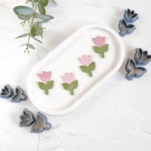 May include: Four pink and green flower-shaped cookie cutters on a white oval plate, and three blue flower-shaped cookie cutters on a white surface.