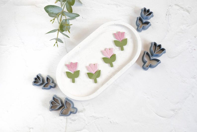 May include: Four pink and green flower-shaped cookie cutters on a white oval plate, and three blue flower-shaped cookie cutters on a white surface.