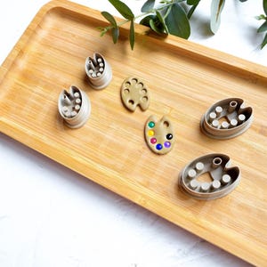 Puede incluir: Un juego de seis cortadores de galletas con forma de paletas de pintura y pinceles. Los cortadores están hechos de plástico gris claro y están dispuestos sobre una superficie de madera.