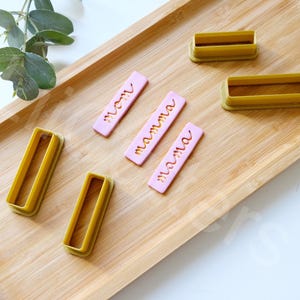 May include: Five cookie cutters on a wooden surface. The cutters are yellow and have the words "mom", "mamma", and "mama" written on them.