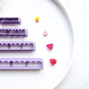 Puede incluir: Cuatro herramientas de plástico morado para hacer rosas de fondant, dispuestas en orden de tamaño, con una muestra de cada tamaño de rosa hecha de fondant en los colores amarillo, rosa, rojo y rojo oscuro.