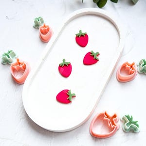 Puede incluir: Un juego de cortadores de galletas de plástico rosa y verde con forma de fresas y zanahorias. Los cortadores están dispuestos sobre una superficie blanca.