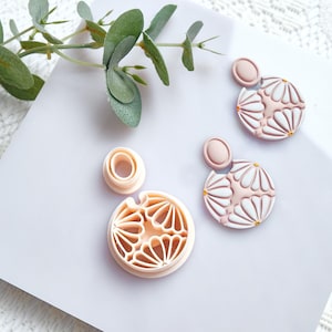 Peut inclure: Un ensemble de boucles d'oreilles en pâte polymère rose clair avec un motif floral, ainsi qu'un emporte-pièce assorti. Les boucles d'oreilles sont de forme circulaire avec un petit dessus ovale. L'emporte-pièce est également circulaire.