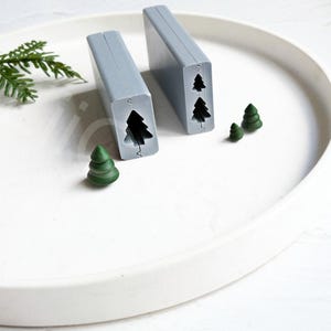 May include: Two gray silicone molds for making small Christmas tree ornaments. The molds are on a white surface with three green Christmas trees made from the molds.