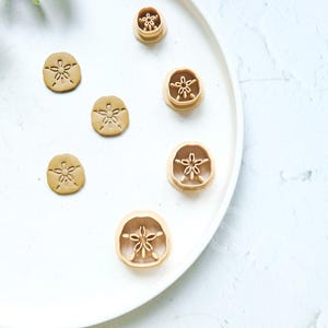 May include: A set of six cookie cutters in the shape of a sand dollar. The cutters are made of metal and have a gold finish. They are arranged on a white plate.