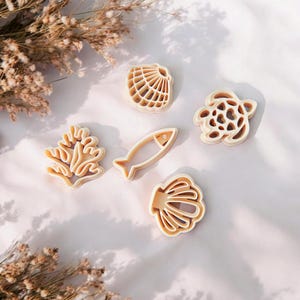 Op de afbeelding: Vijf beige koekjessnijders in de vorm van zeedieren: een schelp, een vis, een schildpad, koraal en nog een schelp. De snijders zijn gerangschikt op een wit oppervlak, met gedroogde bloemen op de achtergrond.