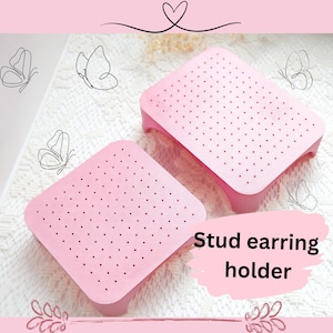 May include: Two pink, square stud earring holders with small holes are displayed. The holders have a raised base and are set against a white, textured background. The words "Stud earring holder" are visible. Delicate butterfly line drawings and a heart are also present.