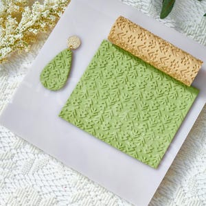 May include: A light green, textured clay earring with a gold-colored stud, a matching green textured square, and a gold-colored textured rolling pin. The earring and square have a repeating geometric pattern. The items are on a white surface.