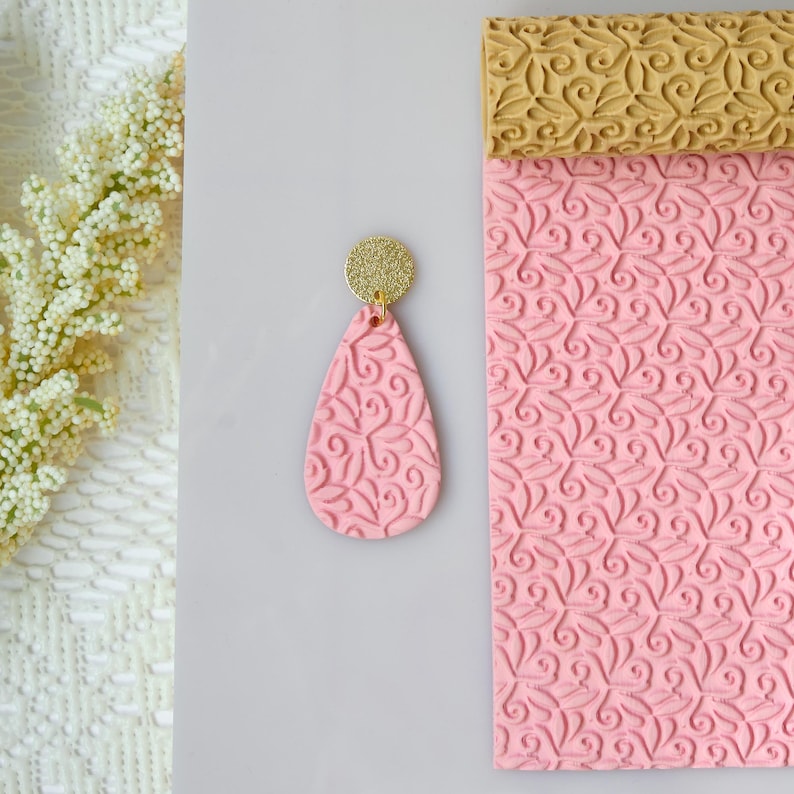 May include: A pink teardrop-shaped earring with a gold glitter disc top. The earring has a detailed embossed floral pattern. A matching pink clay sheet with a floral pattern and a gold clay roller are also visible.