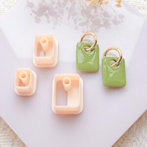 Puede incluir: Vista a&eacute;rea de un conjunto de suministros para manualidades. Se ven tres pendientes rectangulares verde claro con aros dorados y tres cortadores de arcilla rectangulares beige, sobre una superficie blanca.