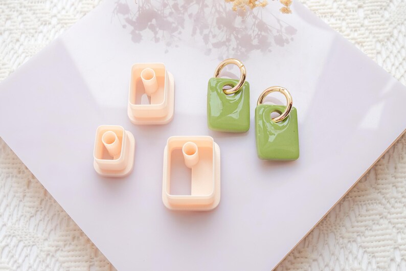 Puede incluir: Vista a&eacute;rea de un conjunto de suministros para manualidades. Se ven tres pendientes rectangulares verde claro con aros dorados y tres cortadores de arcilla rectangulares beige, sobre una superficie blanca.