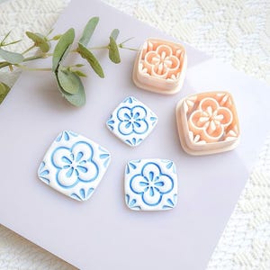 May include: An assortment of decorative ceramic tiles and molds. The tiles feature a white base with blue floral designs. Two molds are peach-colored with a floral pattern. A sprig of greenery is in the upper left corner.