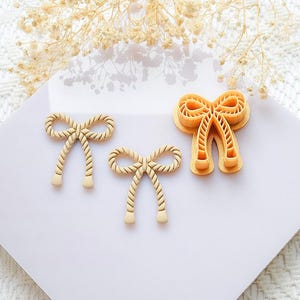 May include: Three beige bow-shaped cookie cutters on a white surface. Two are finished cookies, and one is a cutter. The bows have a rope-like texture. Dried flowers are in the background.