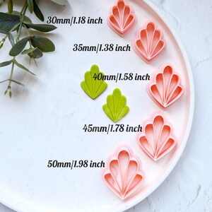 May include: Five different sized pink and green plastic cookie cutters in the shape of a scallop shell. The cutters are arranged in a row on a white plate. The sizes of the cutters are labelled in inches and millimetres.