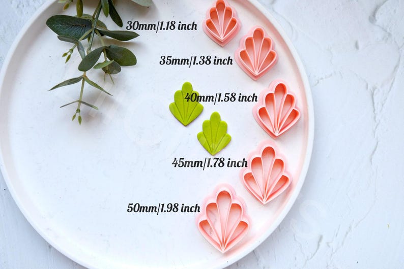 May include: Five different sized pink and green plastic cookie cutters in the shape of a scallop shell. The cutters are arranged in a row on a white plate. The sizes of the cutters are labelled in inches and millimetres.