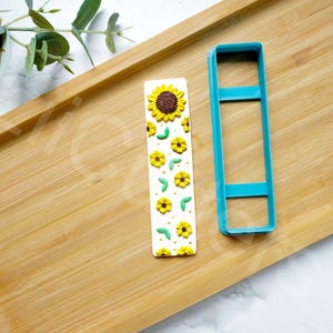 Pode incluir: Um marcador de página branco com um design de girassol e folhas. O marcador de página está ao lado de um cortador de biscoitos de plástico azul.
