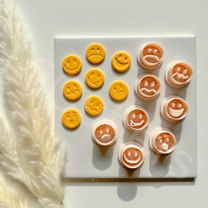 May include: Two sets of cookie cutters in the shape of smiley faces. One set is yellow and the other is peach. The yellow cutters are arranged in a circle, while the peach cutters are stacked on top of each other.