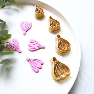 May include: A collection of pink flower-shaped polymer clay pieces and tan flower-shaped cookie cutters arranged on a white plate. Green leafy sprigs are in the upper left corner. The items are for crafting or baking.