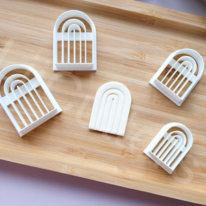 May include: Five white plastic cookie cutters shaped like arches with different numbers of arches. The cutters are on a wooden surface.