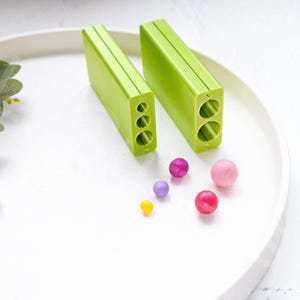 Puede incluir: Dos moldes de plástico verde para hacer caramelos o chocolate redondos. Los moldes tienen agujeros de diferentes tamaños. Hay cinco bolas de caramelo de colores en un plato blanco.