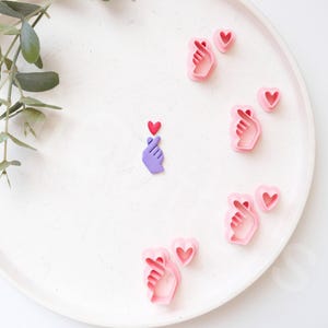 May include: A white plate with pink and purple cookie cutters shaped like hands making a heart sign. The cutters are arranged in a circle around a single purple cutter in the center.