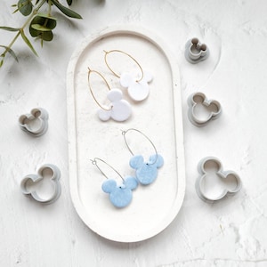 Peut inclure: Deux paires de boucles d'oreilles faites à la main avec un motif Mickey Mouse. Une paire est blanche et l'autre est bleu clair. Les boucles d'oreilles sont présentées sur un plat ovale blanc.