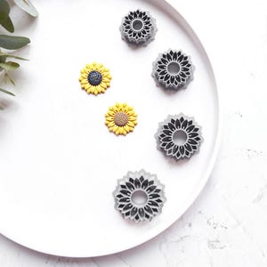 May include: A white plate with five gray and yellow flower-shaped cookie cutters. The cookie cutters are arranged in a circle around the center of the plate. The plate is on a white surface.