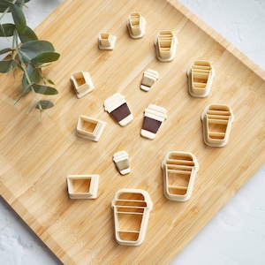 Puede incluir: Un conjunto de 13 cortadores de galletas con forma de tazas de café, sobre una superficie de madera. Los cortadores están hechos de plástico blanco y están dispuestos en una variedad de tamaños y formas.