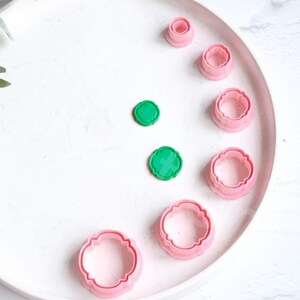 Puede incluir: Un juego de ocho cortadores de galletas de plástico rosa de varios tamaños, con forma de flores, dispuestos en un plato blanco. También se incluyen dos cortadores de galletas de plástico verde.