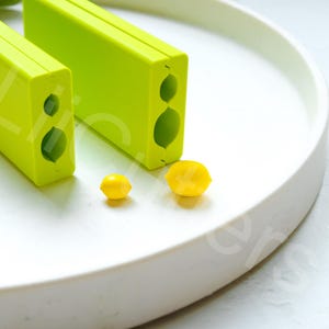 Könnte beinhalten: Zwei rechteckige, limegrüne Kunststoffformen mit abgerundeter Kante. Die Formen haben zwei unterschiedlich große runde Ausschnitte. Zwei kleine, gelbe, zitronenförmige Bonbons liegen vor den Formen.