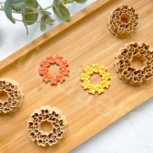 Puede incluir: Una bandeja de madera con varios cortadores de galletas en forma de corona en tonos beige, naranja y amarillo. Hojas verdes en la esquina superior izquierda. Los cortadores son probablemente para hornear o manualidades.