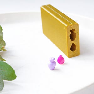 Op de afbeelding: Een gouden plastic mal met twee vaasvormige uitsnijdingen. Twee kleine, kleurrijke, decoratieve vazen staan naast de mal.