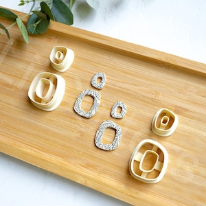 May include: A set of seven white plastic cookie cutters in various shapes and sizes, including a rectangle, a square, and a circle. The cutters are arranged on a wooden surface.
