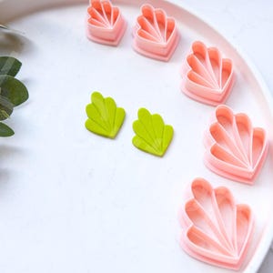 May include: A set of pink and green plastic cookie cutters shaped like seashells. The cutters are arranged on a white plate.