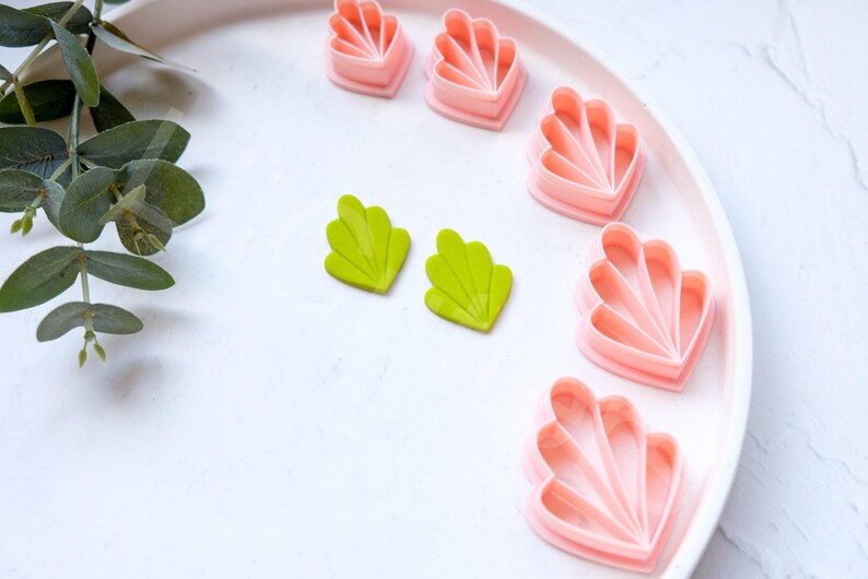 May include: A set of pink and green plastic cookie cutters shaped like seashells. The cutters are arranged on a white plate.