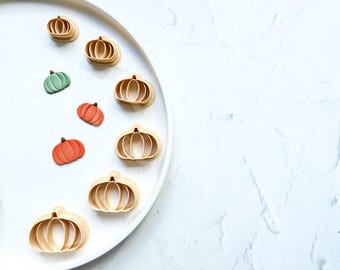 May include: A white plate with eight pumpkin-shaped cookie cutters and three small orange and green pumpkins. The cookie cutters are made of wood and have a natural finish.